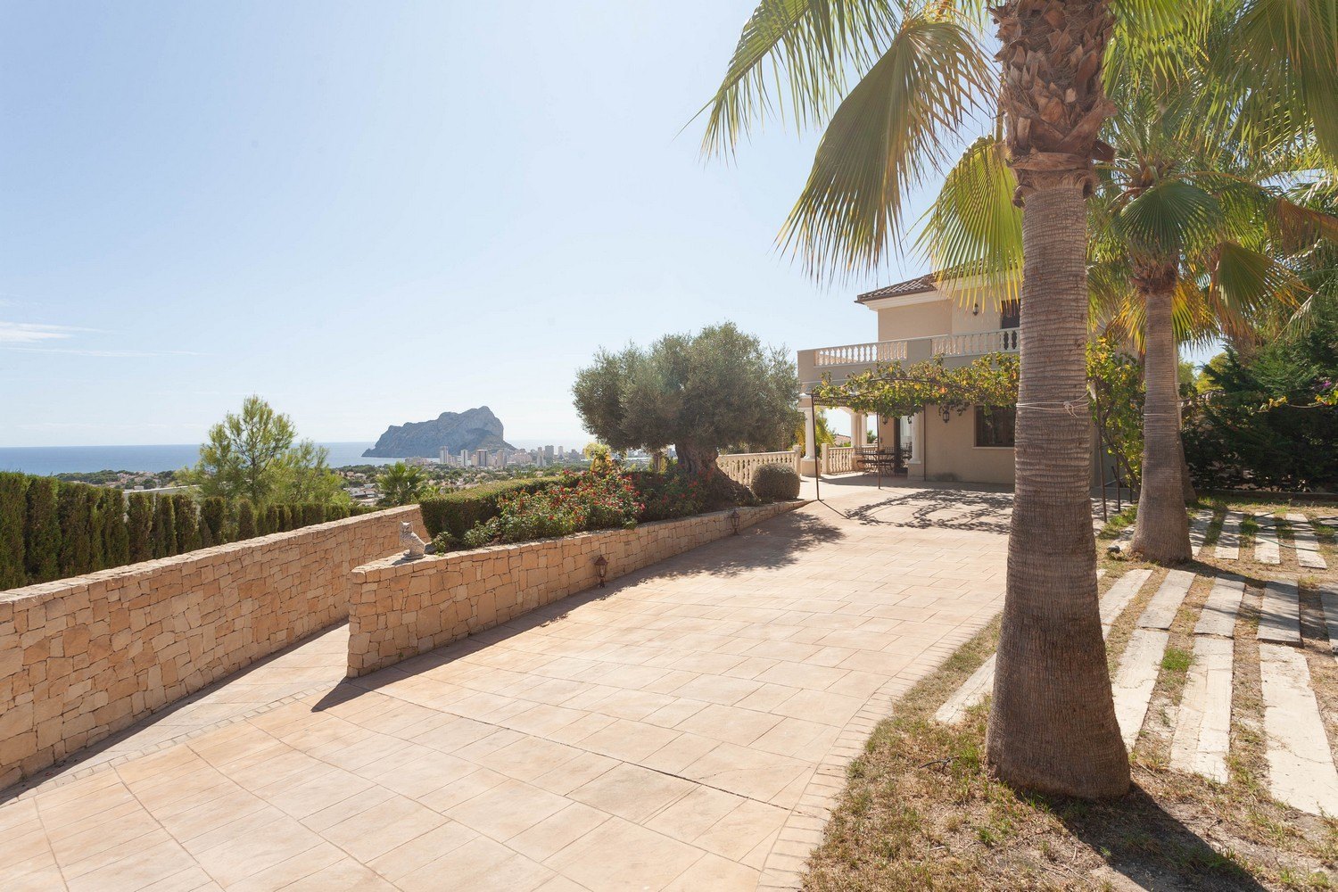 Vue extérieure de la villa avec allée carrelée, mur en pierre et vue sur la mer.