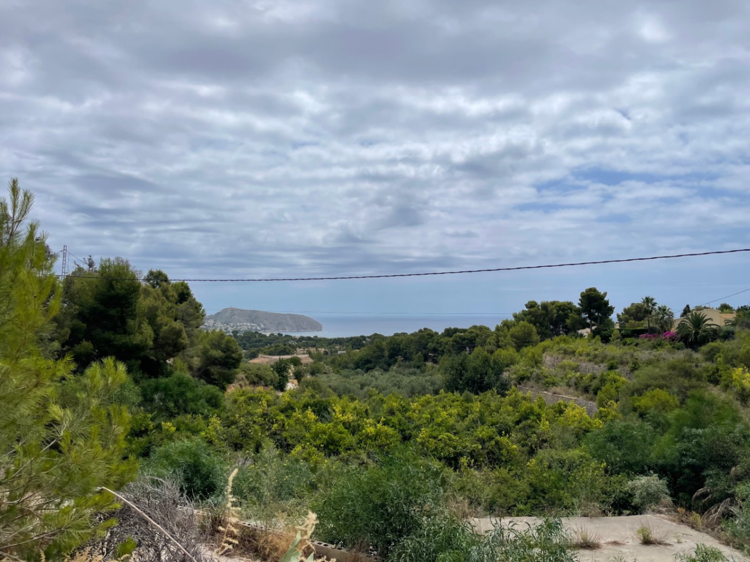 Vue panoramique depuis le terrain vers la côte de Moraira, avec le Cap d'Or et la mer Méditerranée se profilant derrière une végétation dense.