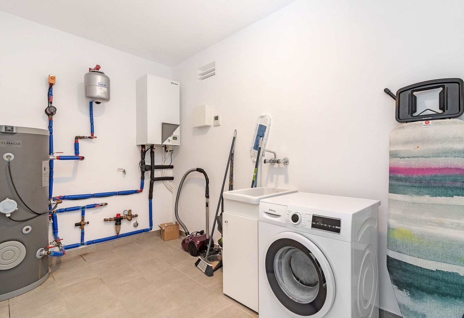 Buanderie et local technique avec lave-linge, évier, système aérothermique Daikin et réservoir d'eau sur carrelage gris.