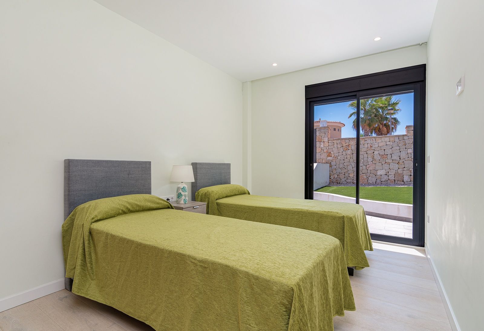 Chambre avec deux lits simples, têtes de lit grises et porte coulissante en verre offrant un accès direct au jardin et à la terrasse.