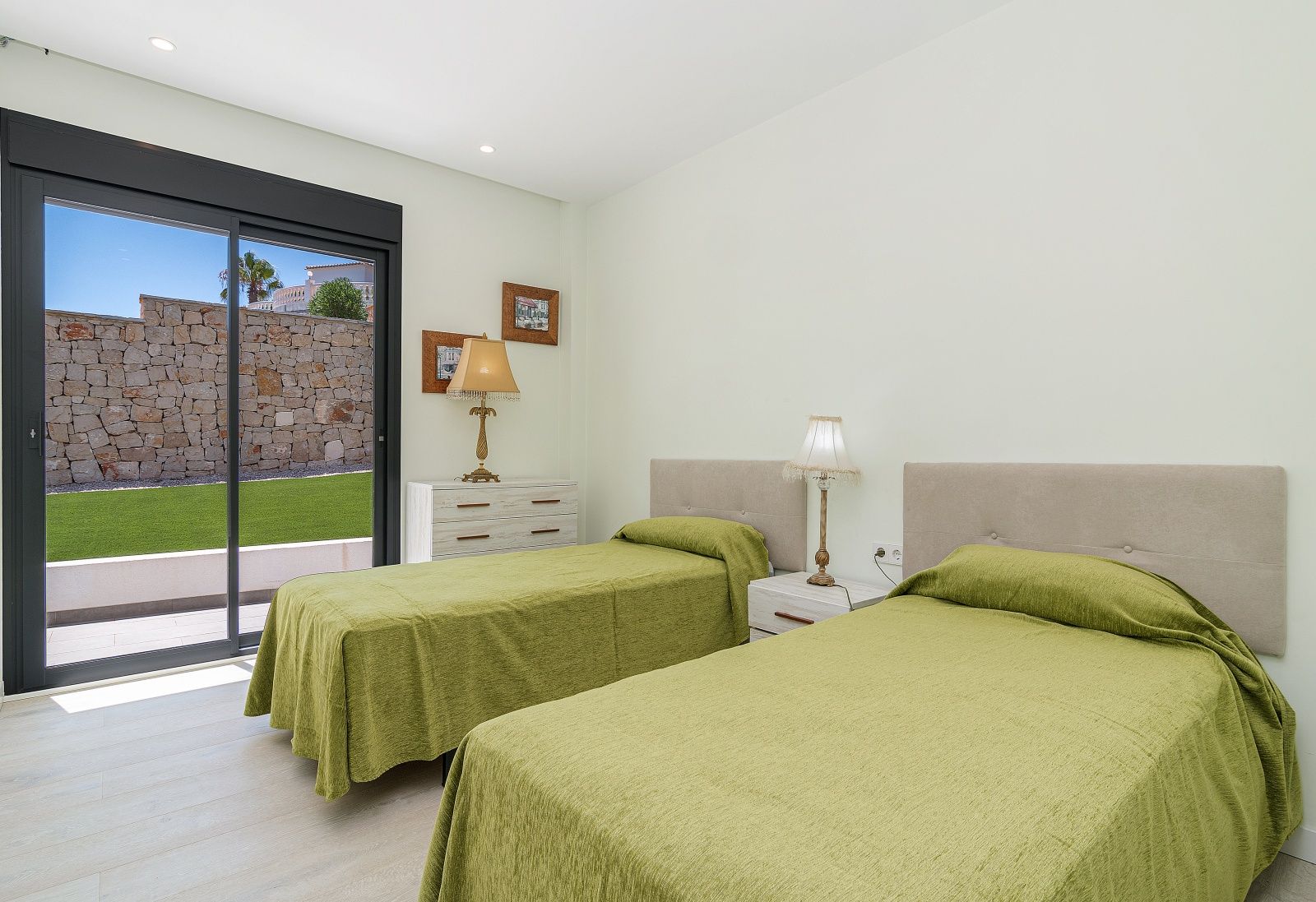 Chambre avec deux lits simples, têtes de lit rembourrées et accès extérieur vers le jardin. Sol en grès cérame aspect bois et éclairage encastré au plafond.