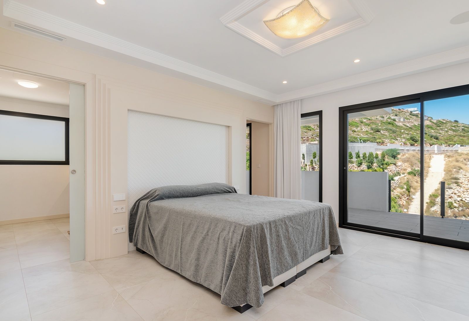 Chambre lumineuse avec accès terrasse par baies vitrées, niche de tête de lit intégrée et éclairage encastré au plafond avec moulures élégantes.