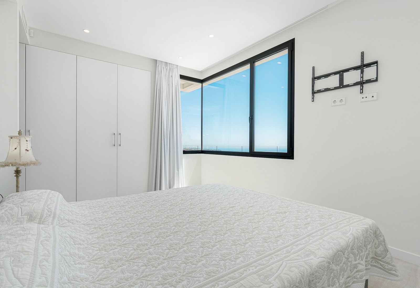 Chambre lumineuse avec placards intégrés blancs, éclairage encastré et fenêtre d'angle aux cadres noirs offrant une vue dégagée sur la mer Méditerranée.