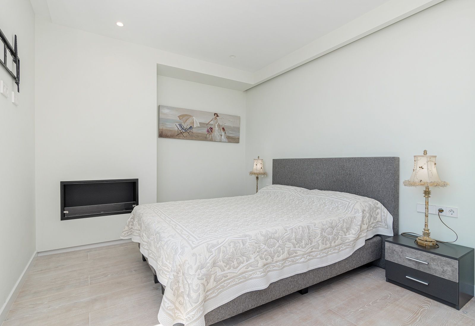 Chambre moderne avec cheminée murale intégrée, sol en grès cérame imitation bois et tête de lit rembourrée grise.