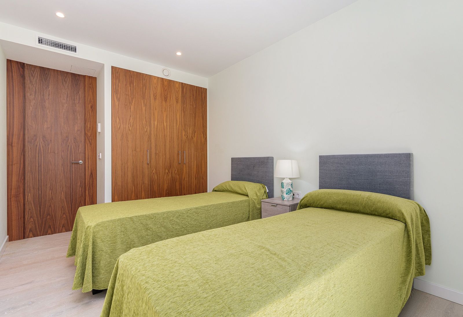 Chambre moderne avec deux lits simples, placards intégrés en bois naturel, climatisation gainable et éclairage encastré au plafond.