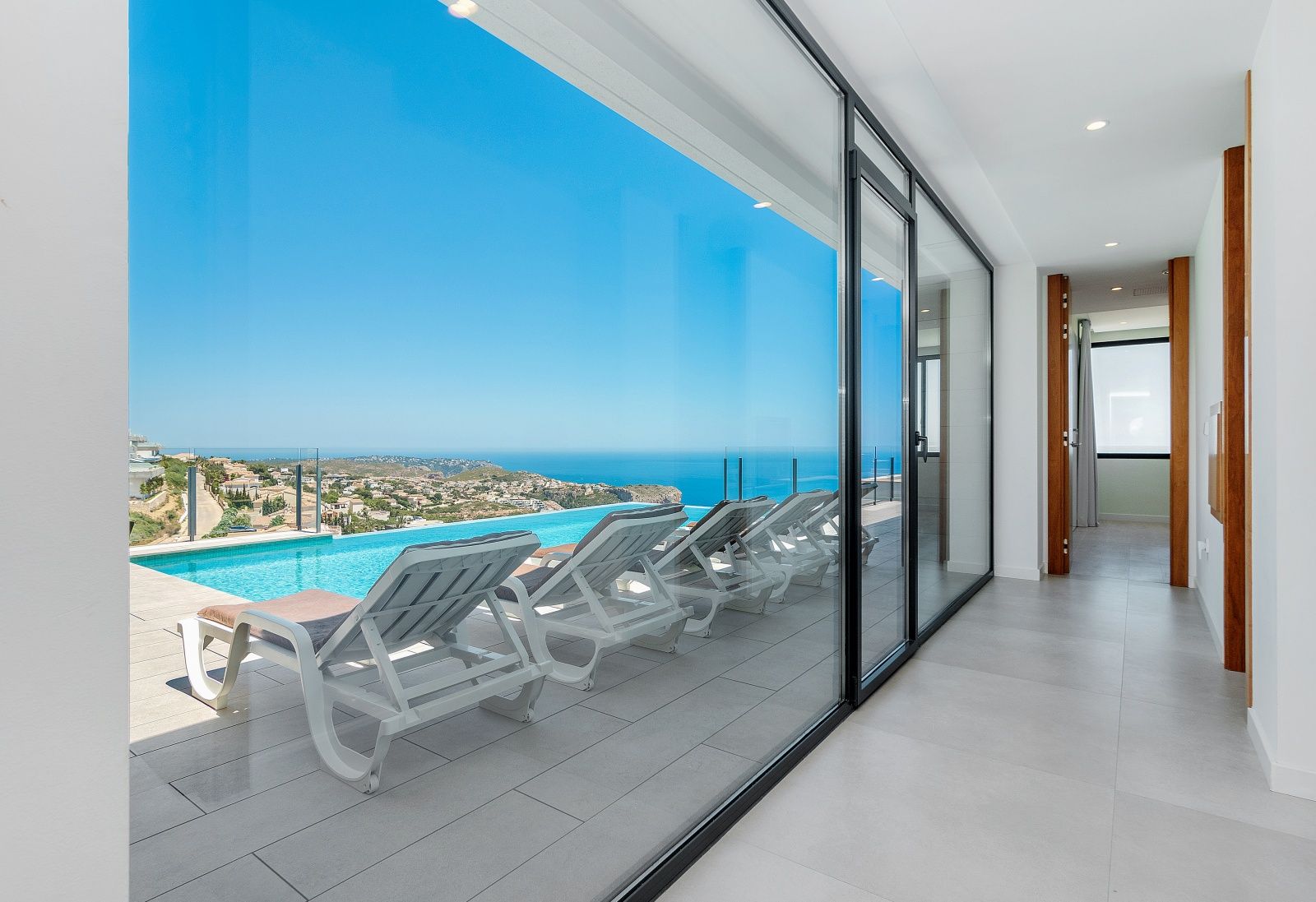 Couloir intérieur avec de grandes baies vitrées coulissantes ouvrant sur la terrasse de la piscine à débordement et offrant une vue panoramique sur la mer.