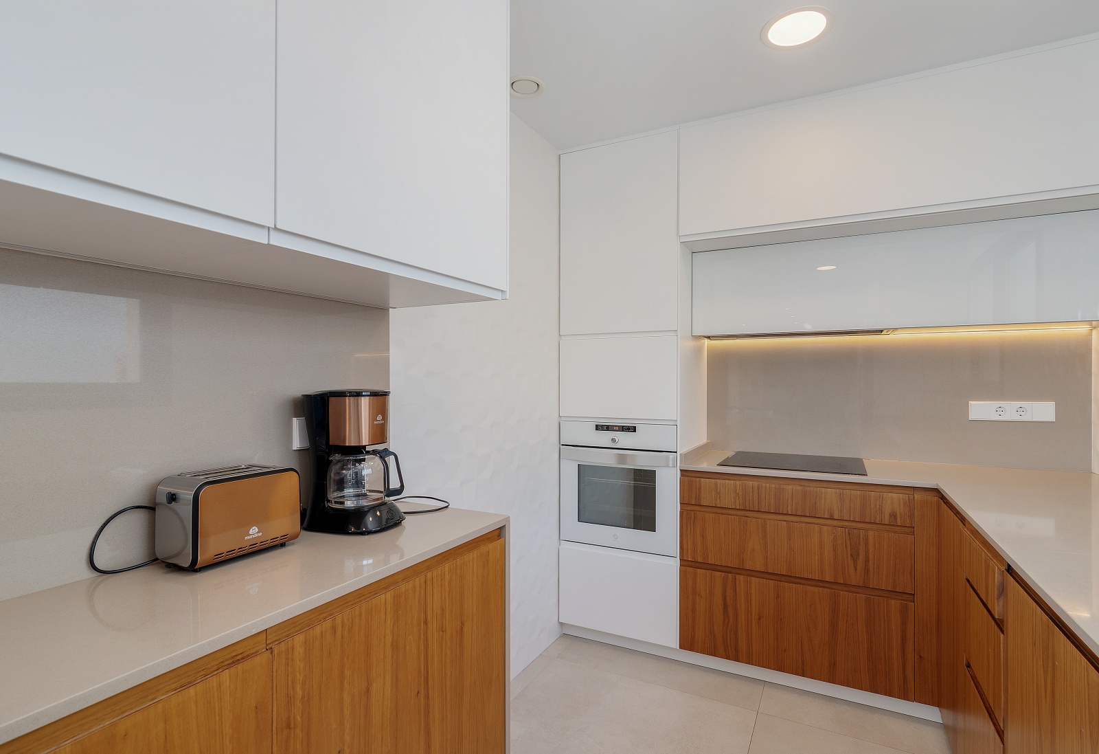 Cuisine moderne avec placards blancs et bois, équipée d'un four encastré, d'une plaque à induction et d'un plan de travail en quartz avec éclairage LED.