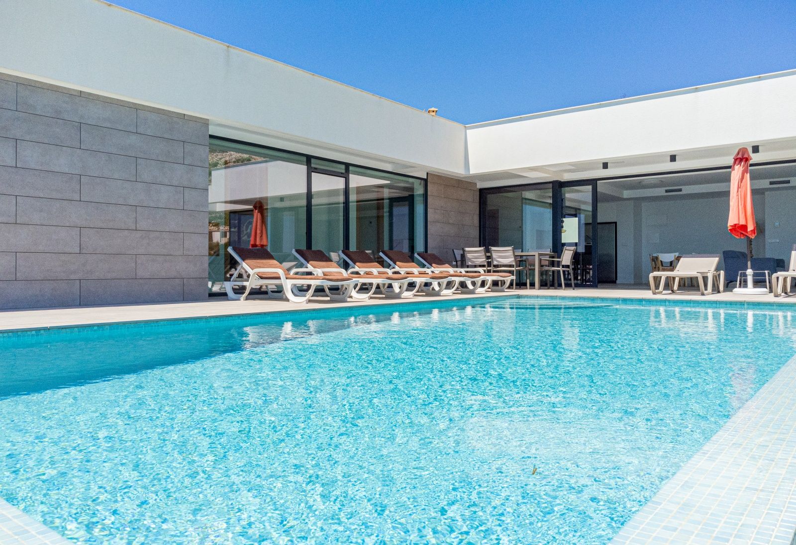 Espace piscine privée avec terrasse ensoleillée, transats, coin repas extérieur et architecture moderne aux larges baies vitrées sous un ciel bleu azur.