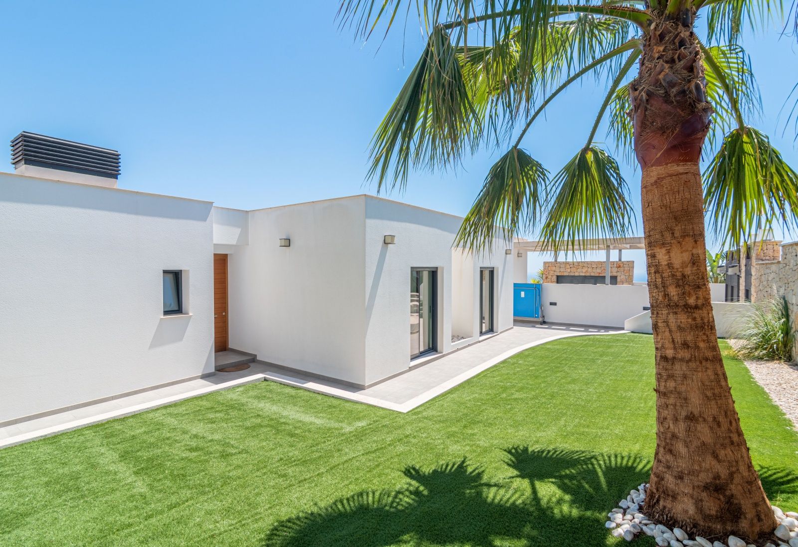 Extérieur d'une villa moderne avec façade blanche, pelouse synthétique et palmier. L'image montre la porte d'entrée en bois et de larges baies vitrées.