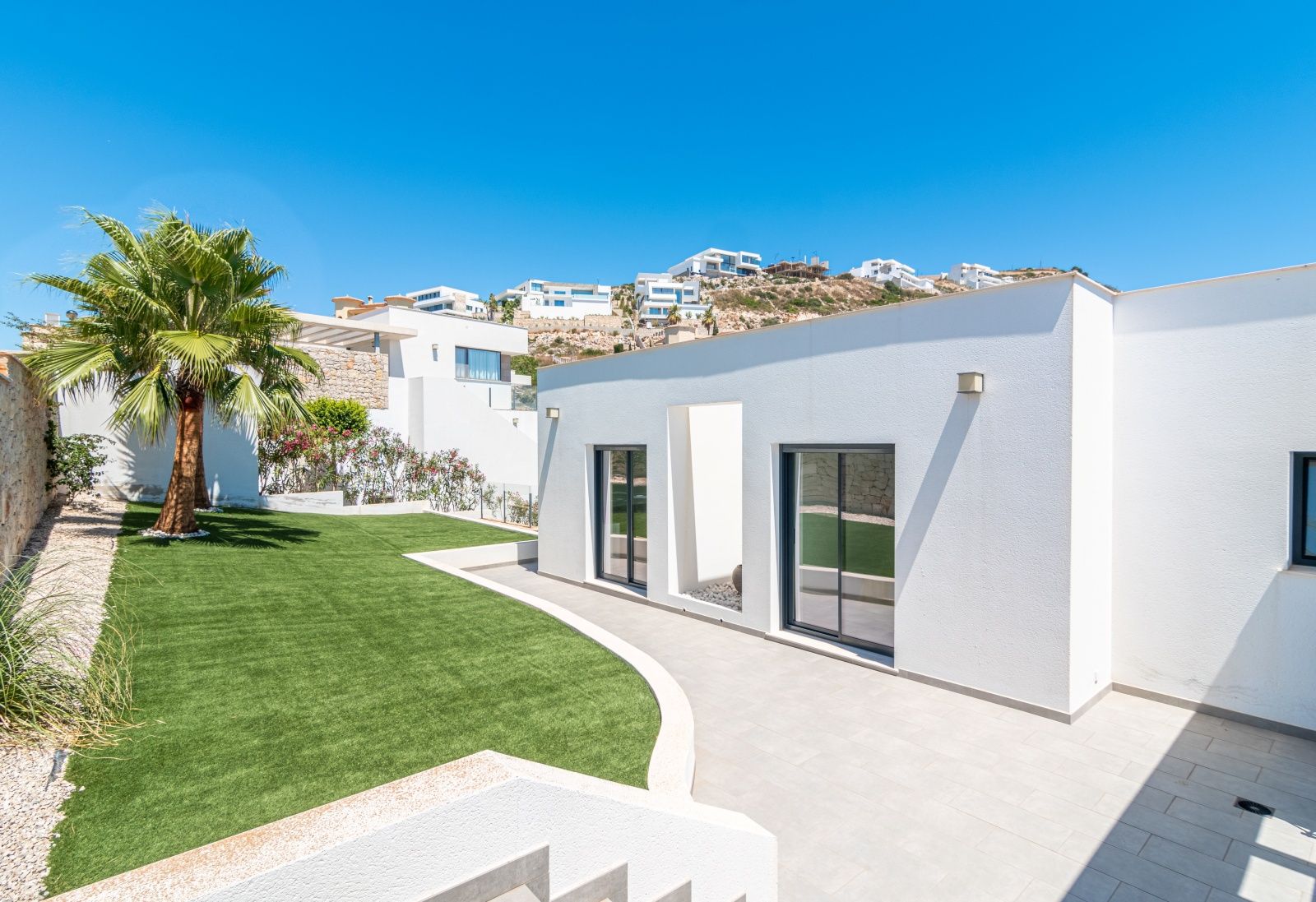 Façade moderne blanche avec de grandes baies vitrées, un jardin de pelouse synthétique agrémenté d'un palmier et une terrasse pavée élégante.