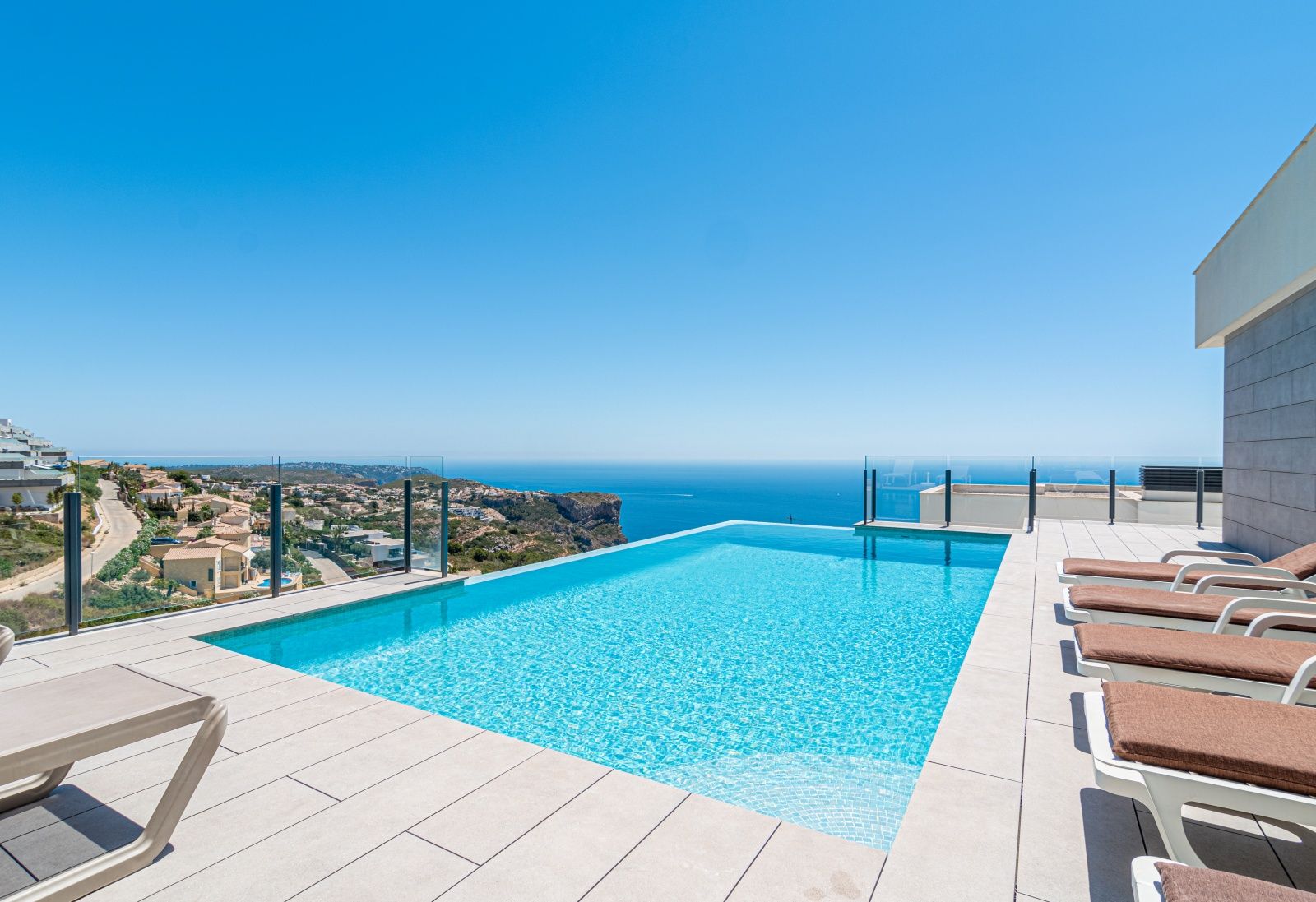 Piscine à débordement avec vue panoramique sur la mer, entourée d'une terrasse en carrelage grand format et de garde-corps en verre.