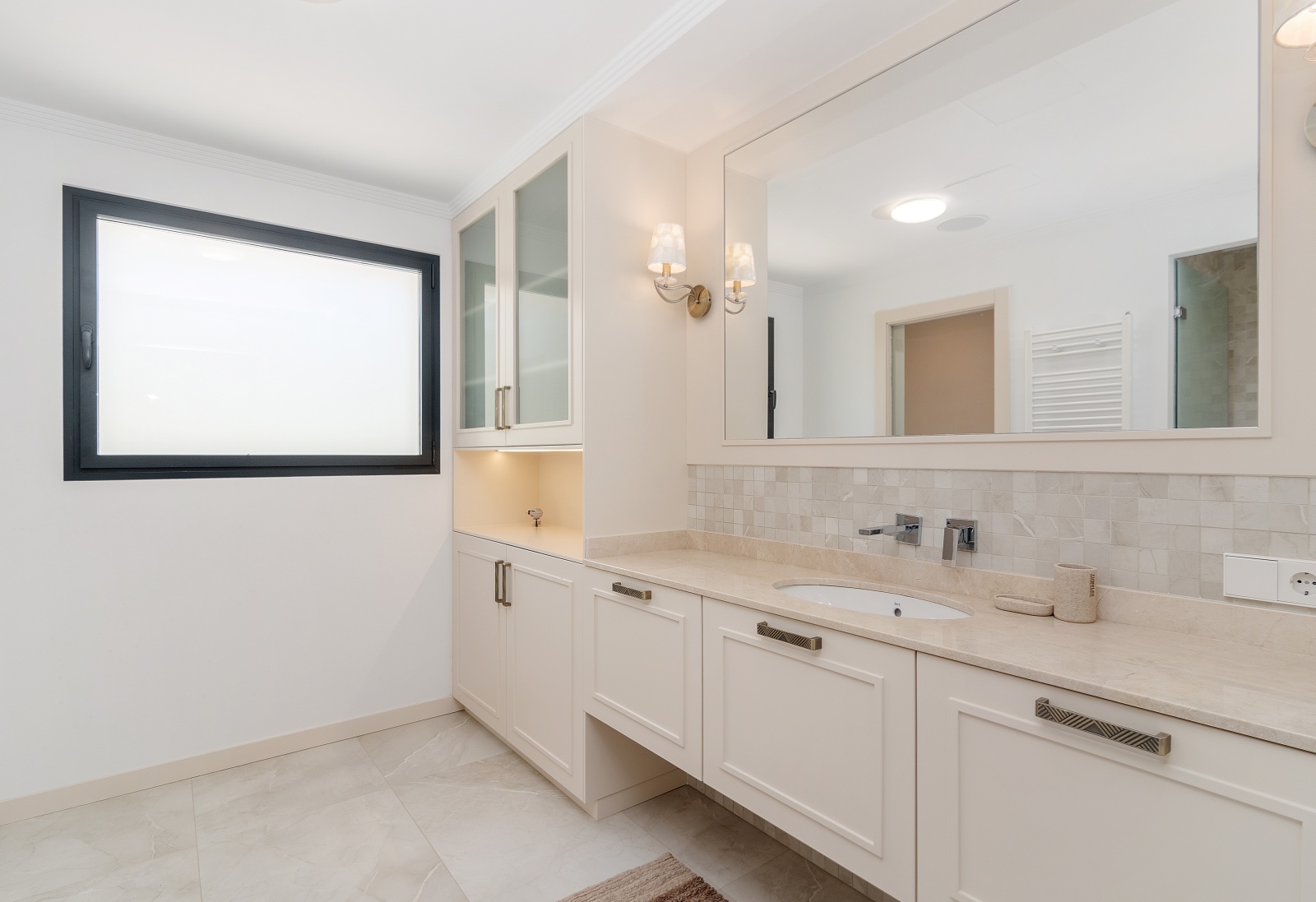 Salle de bain élégante avec plan de travail en pierre, vasque intégrée et meubles crème. Comprend des vitrines éclairées et des appliques murales classiques près du miroir.
