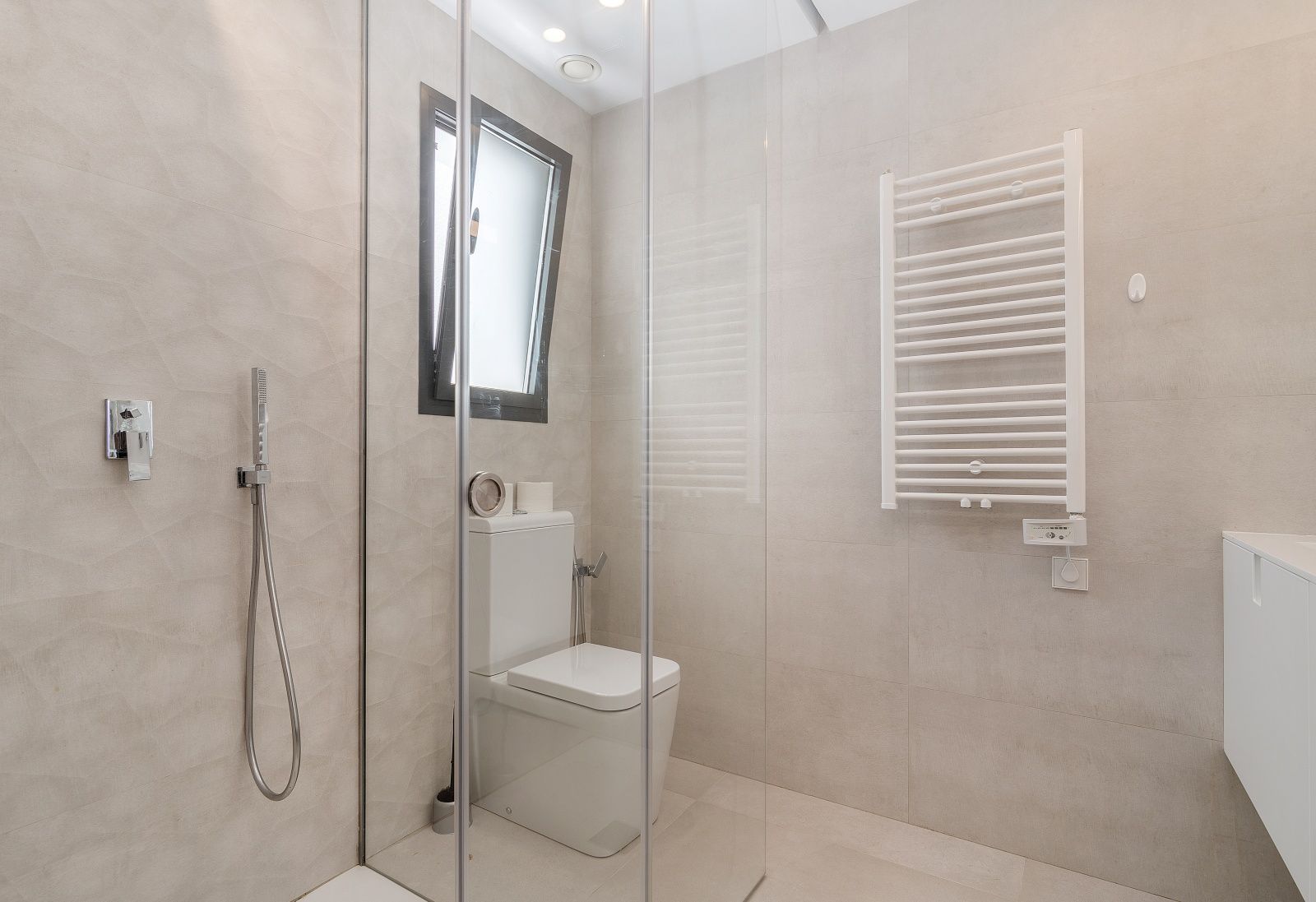 Salle de bain moderne avec douche à l'italienne vitrée, carrelage mural beige texturé et radiateur sèche-serviettes blanc fixé au mur.