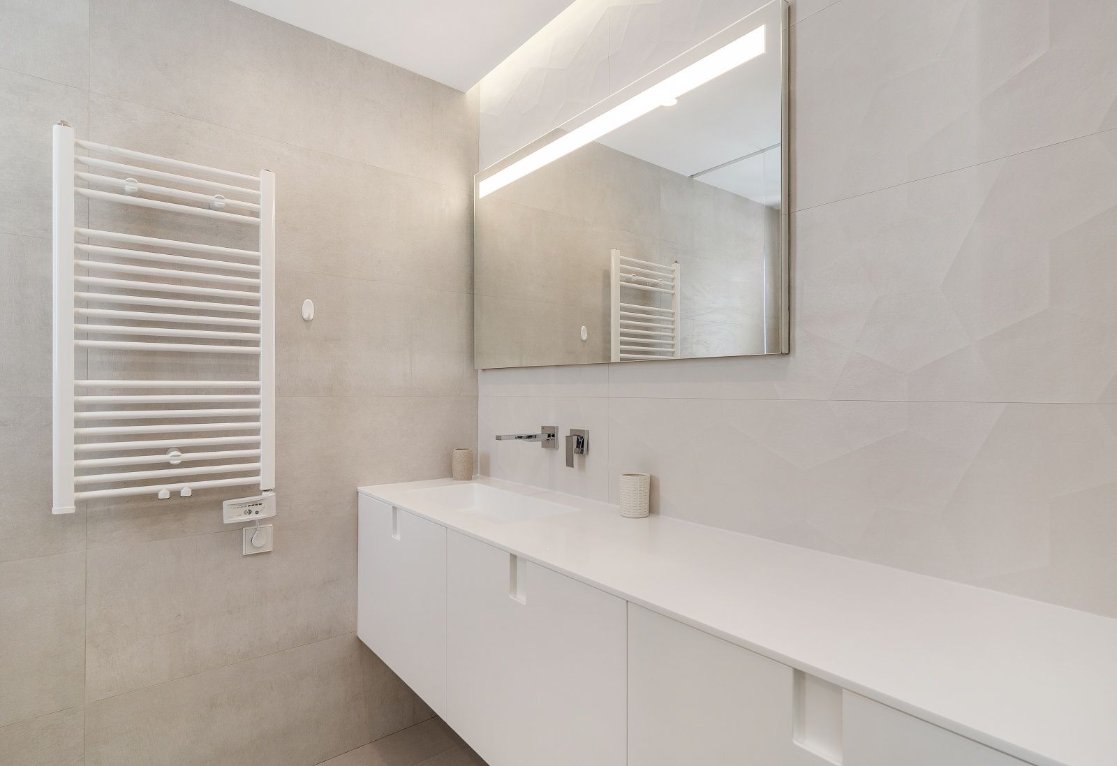 Salle de bain moderne avec meuble vasque suspendu blanc, robinetterie encastrée, miroir rétroéclairé par LED et radiateur sèche-serviettes blanc.