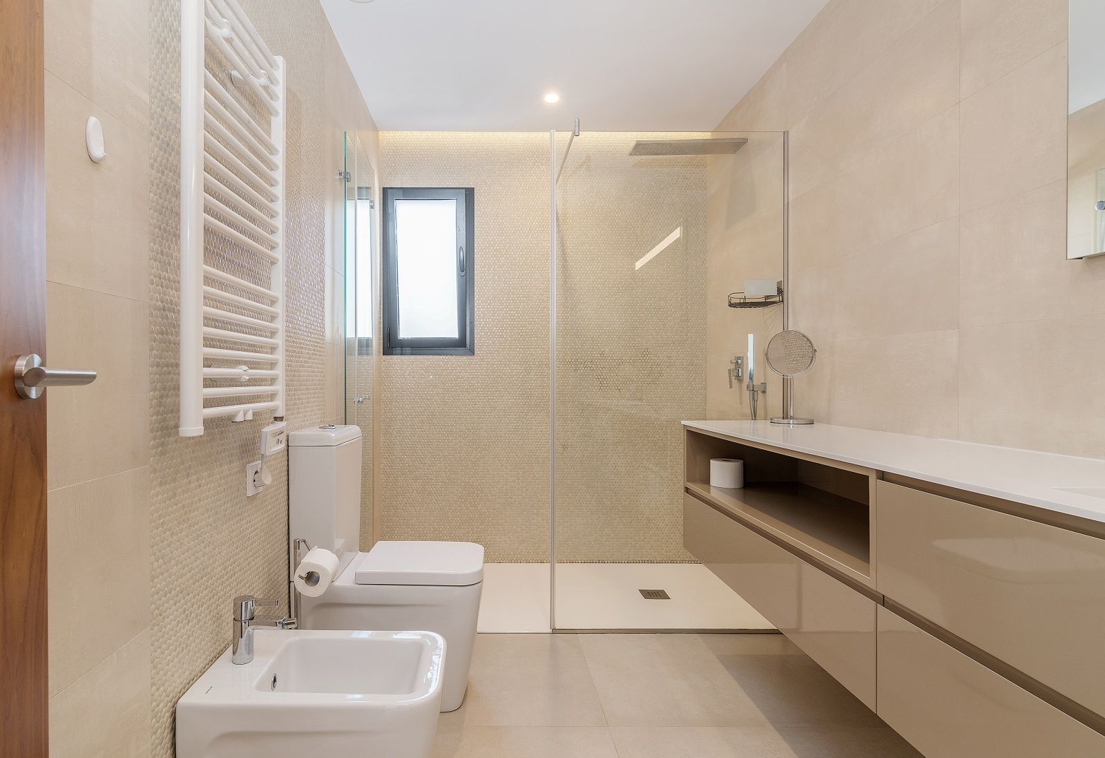 Salle de bains moderne avec douche à l'italienne, paroi en verre, meuble vasque suspendu, bidet et sèche-serviettes blanc sur carrelage beige.