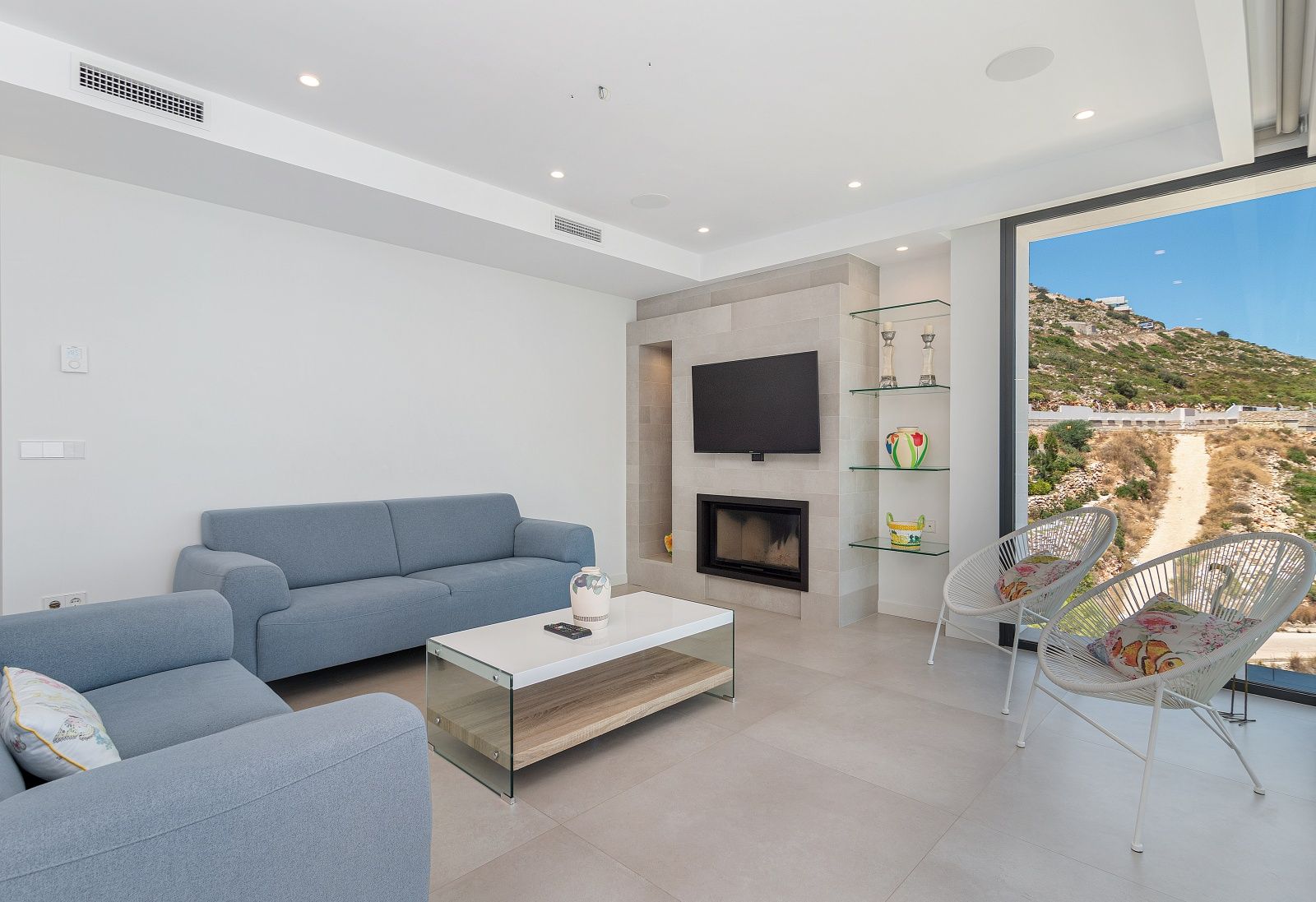 Salon contemporain avec canapés bleus, cheminée intégrée dans un mur en pierre, étagères en verre et grandes ouvertures sur le paysage naturel.