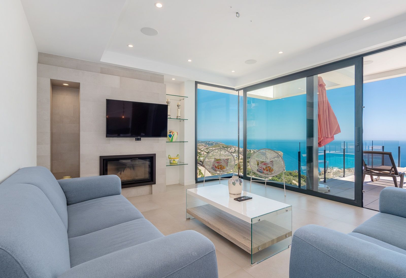 Salon moderne avec canapés bleus, cheminée intégrée et grandes baies vitrées offrant une vue panoramique sur la mer Méditerranée et le littoral.