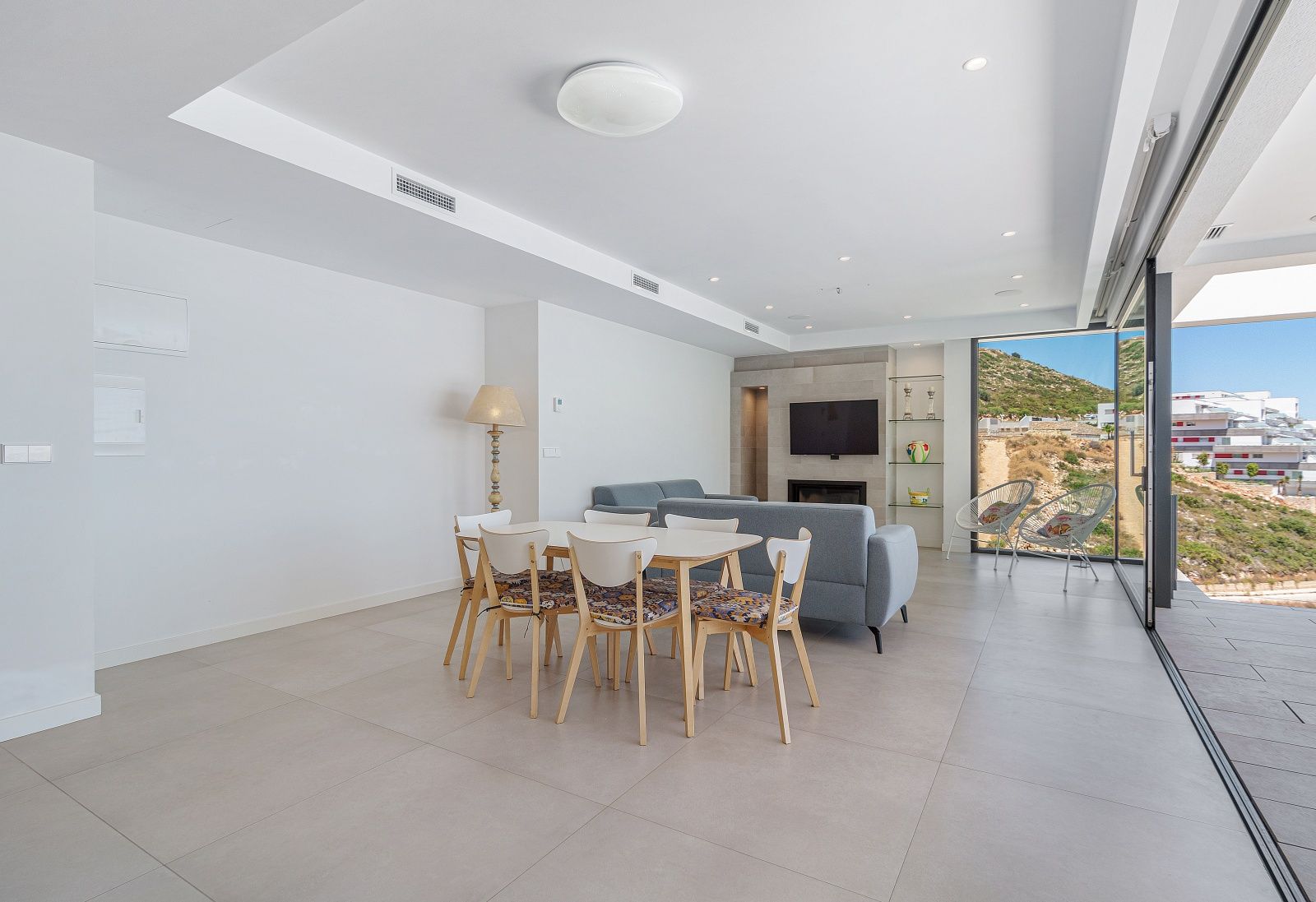 Salon-salle à manger lumineux avec carrelage grand format, mur TV avec cheminée intégrée et larges baies vitrées coulissantes ouvrant sur l'extérieur.