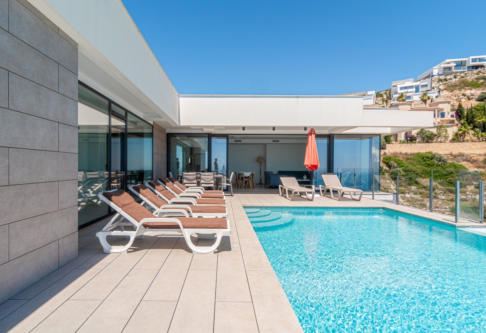 Terrasse principale avec piscine à débordement, transats et coin repas extérieur. Façade moderne avec de larges baies vitrées et des panneaux en pierre grise.