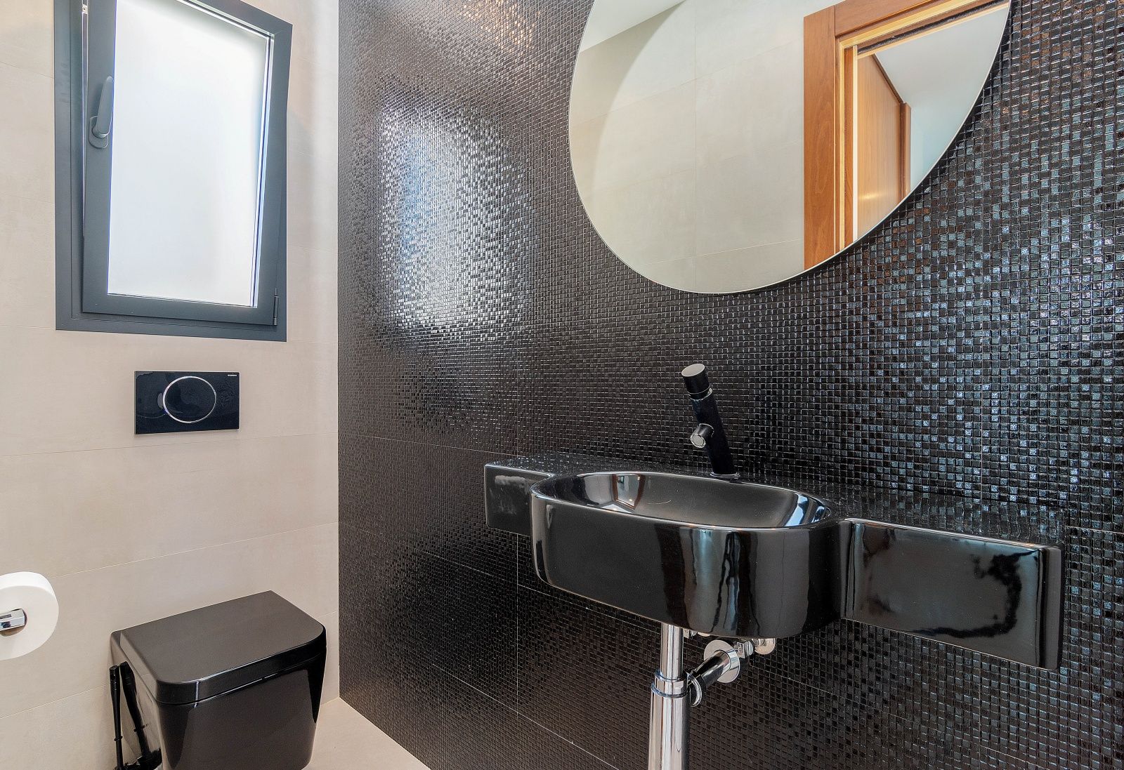 Toilettes d'invités modernes avec mur en mosaïque noire, lavabo et WC suspendus noirs, miroir circulaire et fenêtre latérale.