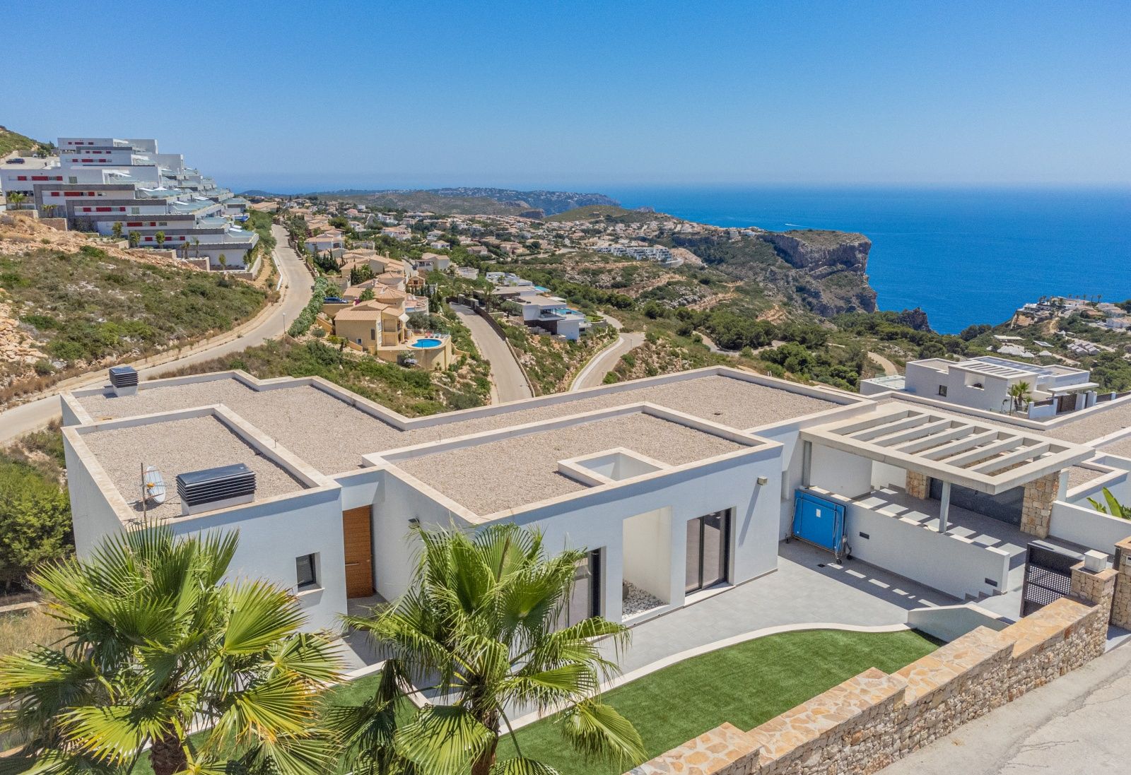 Vue aérienne de la villa moderne à Cumbre del Sol, avec ses toits plats, ses façades blanches et une vue panoramique sur la mer Méditerranée et les falaises.