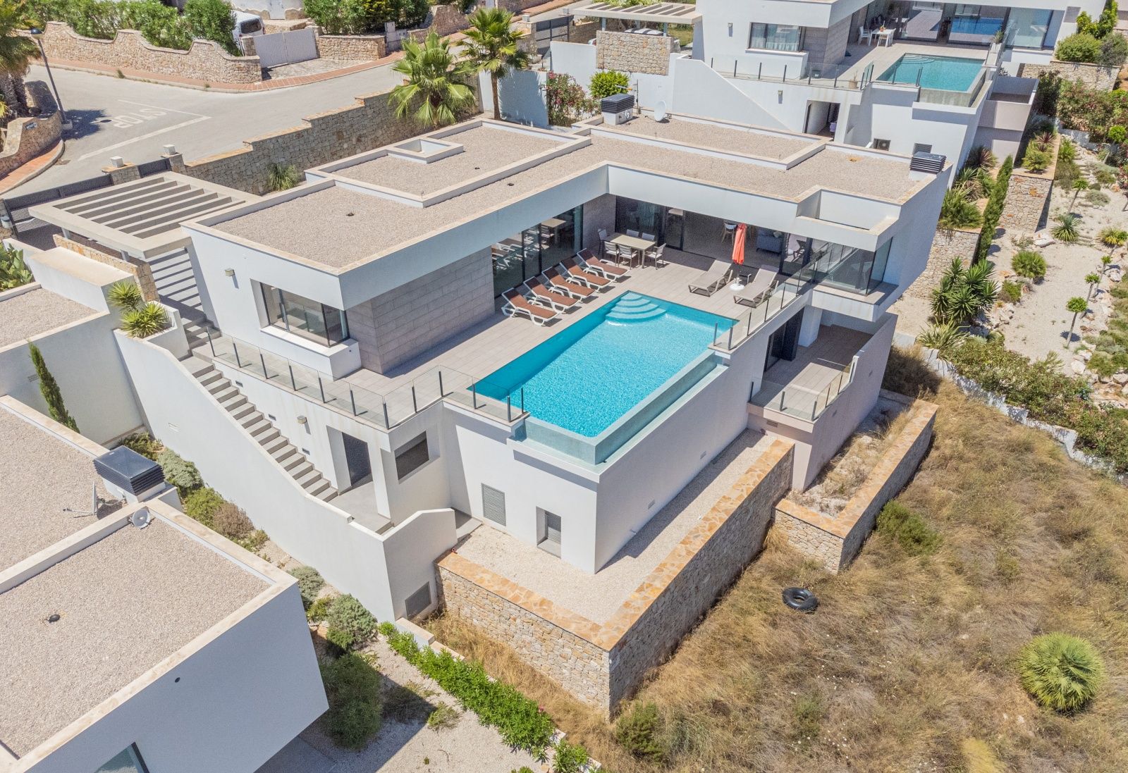 Vue aérienne de la villa moderne avec piscine à débordement et paroi en verre, grande terrasse ensoleillée et architecture épurée aux murs blancs.