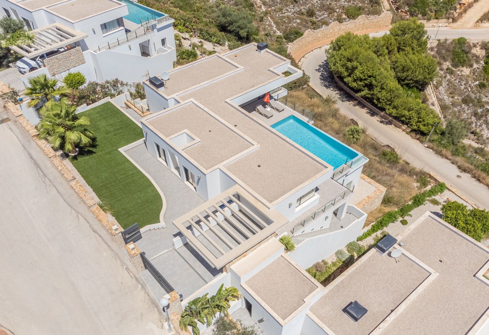 Vue aérienne de la villa moderne avec piscine à débordement, toits plats en gravier, pelouse synthétique et parking couvert par une pergola.