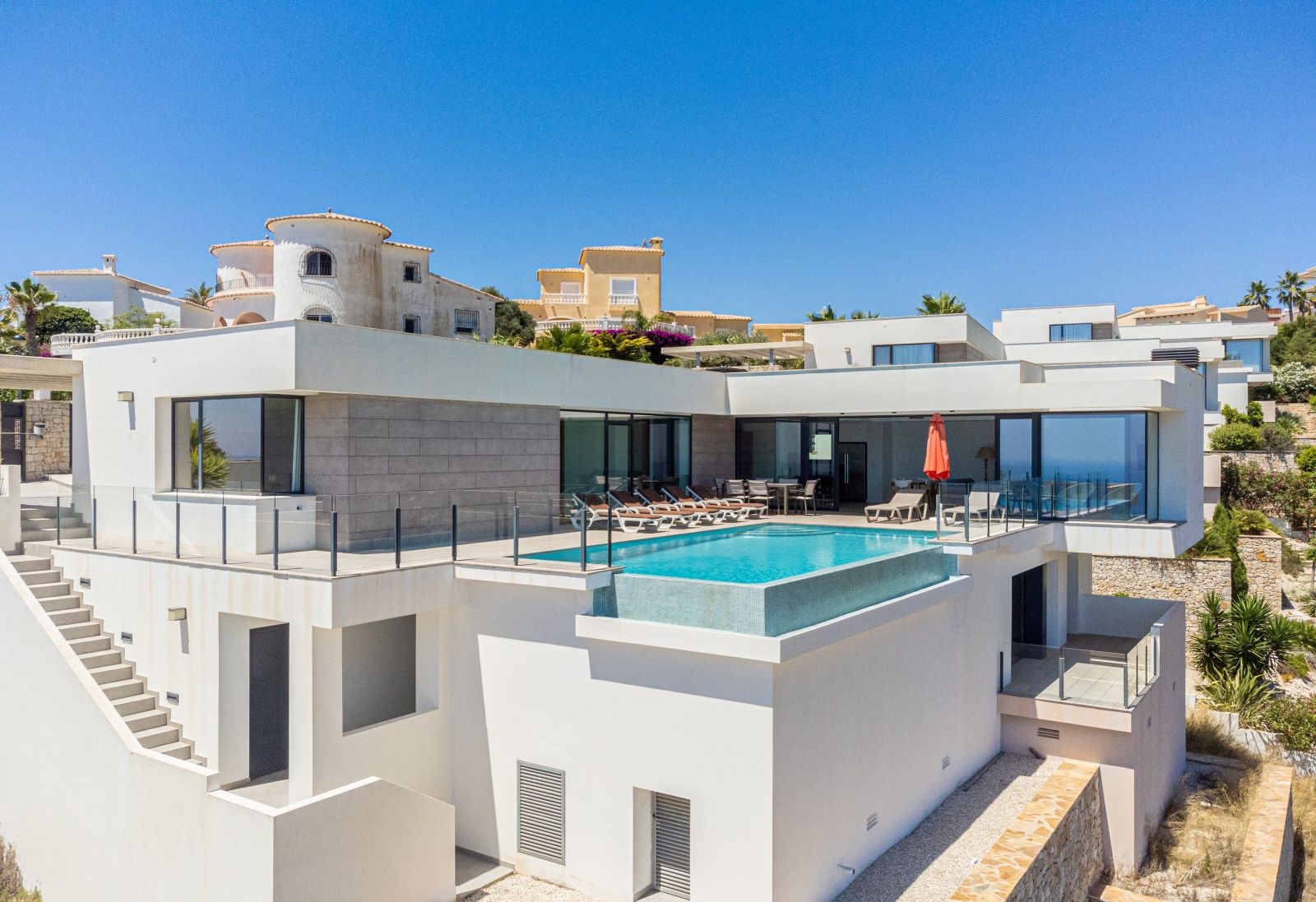 Vue extérieure de la villa moderne avec piscine à débordement, grande terrasse ensoleillée et façade blanche aux accents de pierre grise.