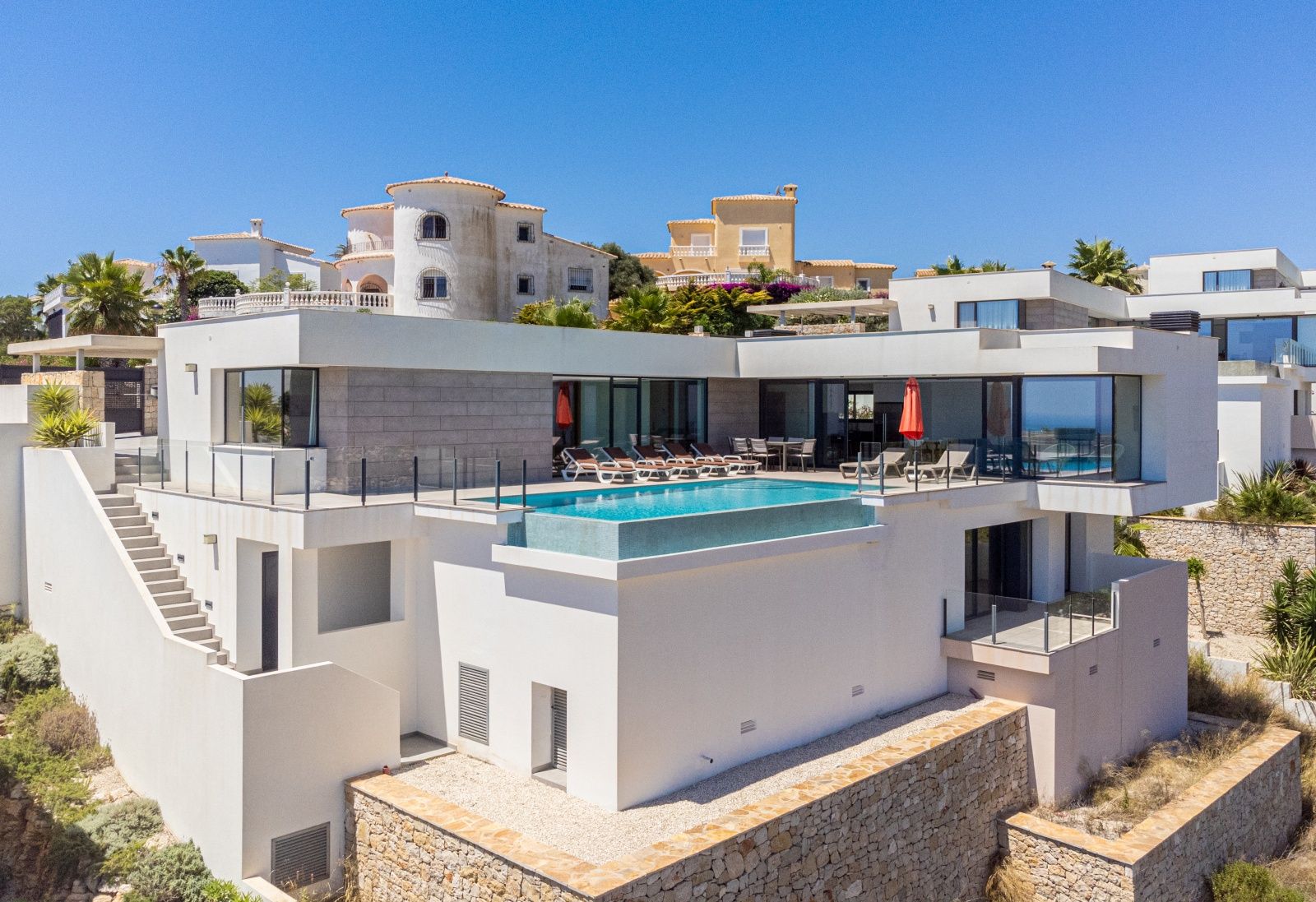 Vue extérieure de la villa moderne avec piscine à débordement, grandes terrasses avec garde-corps en verre et façade blanche aux détails en pierre.