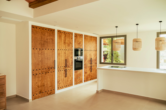 Cuisine de design avec placards en bois rustique et électroménagers intégrés. Un îlot blanc avec plaque à induction est éclairé par trois suspensions en fibres naturelles.