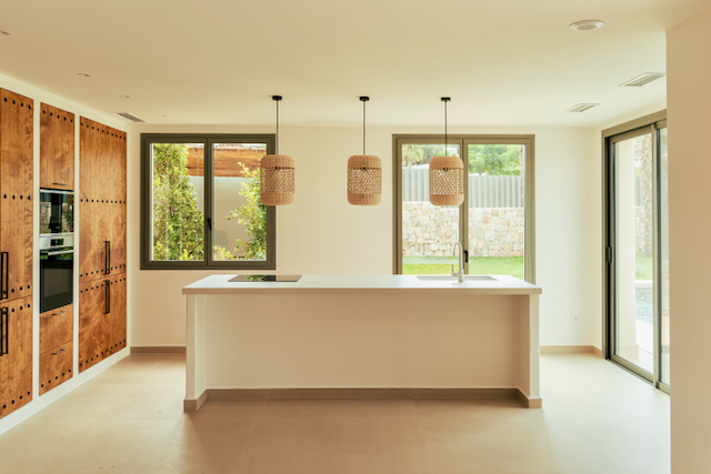 Cuisine ouverte avec îlot central blanc, suspensions en fibres naturelles et placards en bois. Les larges baies vitrées offrent une vue dégagée sur le jardin et la piscine.