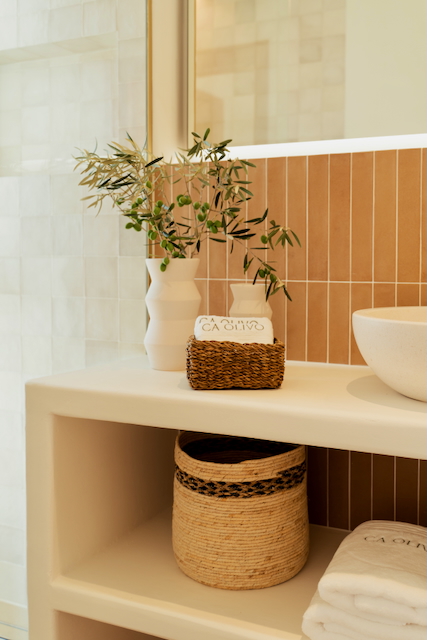 Détail de la salle de bain avec plan de travail en microciment, carrelage vertical terre cuite et étagères ouvertes. Décoration naturelle avec paniers en osier et branches d'olivier.