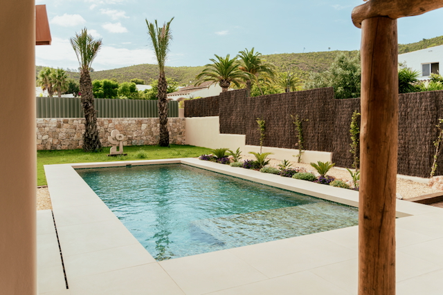 Piscine rectangulaire avec terrasse en grès cérame clair, jardin paysager avec palmiers, mur en pierre naturelle et vue sur les collines environnantes.