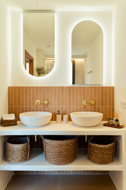 Salle de bain avec double vasque en pierre, robinetterie dorée encastrée et carrelage vertical terracotta. Miroirs cintrés rétroéclairés et paniers en osier pour le rangement.