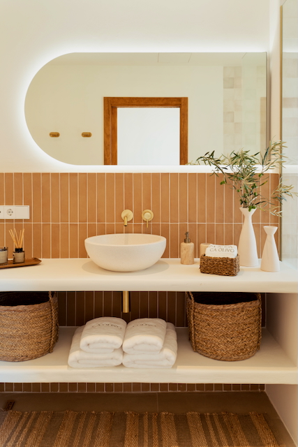 Salle de bain moderne avec vasque à poser, robinetterie en laiton et carrelage vertical terracotta. Un miroir ovale rétroéclairé surplombe un plan de toilette aux rangements ouverts.