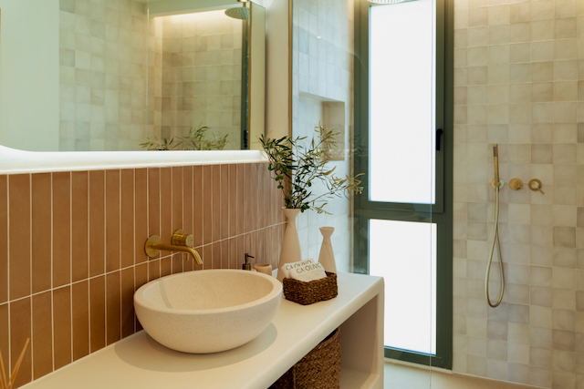 Salle de bains moderne avec vasque en pierre, robinetterie dorée brossée et carrelage vertical terre cuite. Comprend une douche vitrée et une belle luminosité naturelle.
