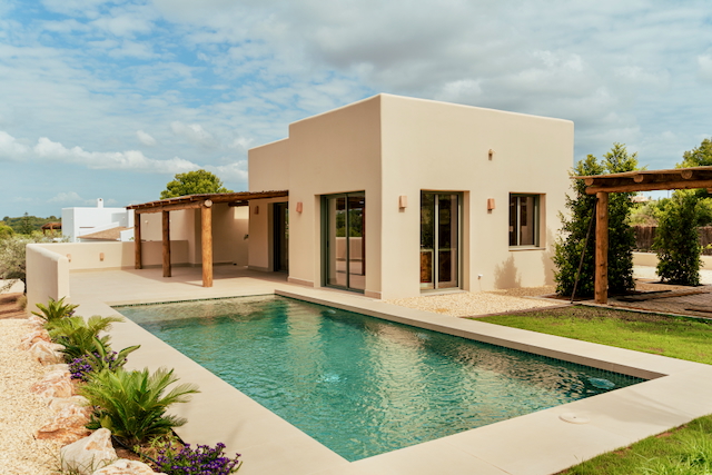 Vue extérieure de la villa moderne avec piscine rectangulaire, façade aux tons sable et pergola en bois rustique intégrée à la grande terrasse.