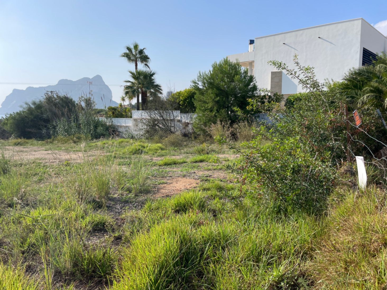 Terrain constructible plat à Bassetes, Calpe, offrant une vue sur le Peñón de Ifach. La parcelle est entourée de végétation locale et d'une villa moderne.