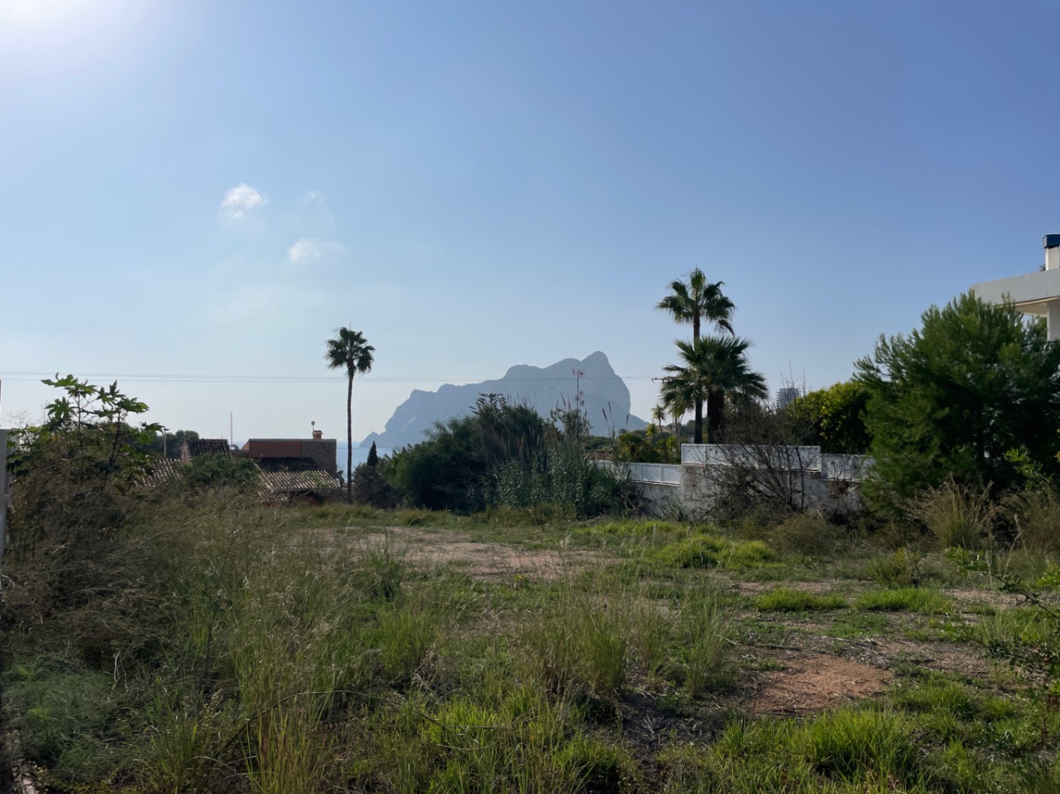 Terrain plat à Bassetes offrant une vue sur le Peñón de Ifach, entouré de végétation méditerranéenne et de palmiers sous un ciel azur.