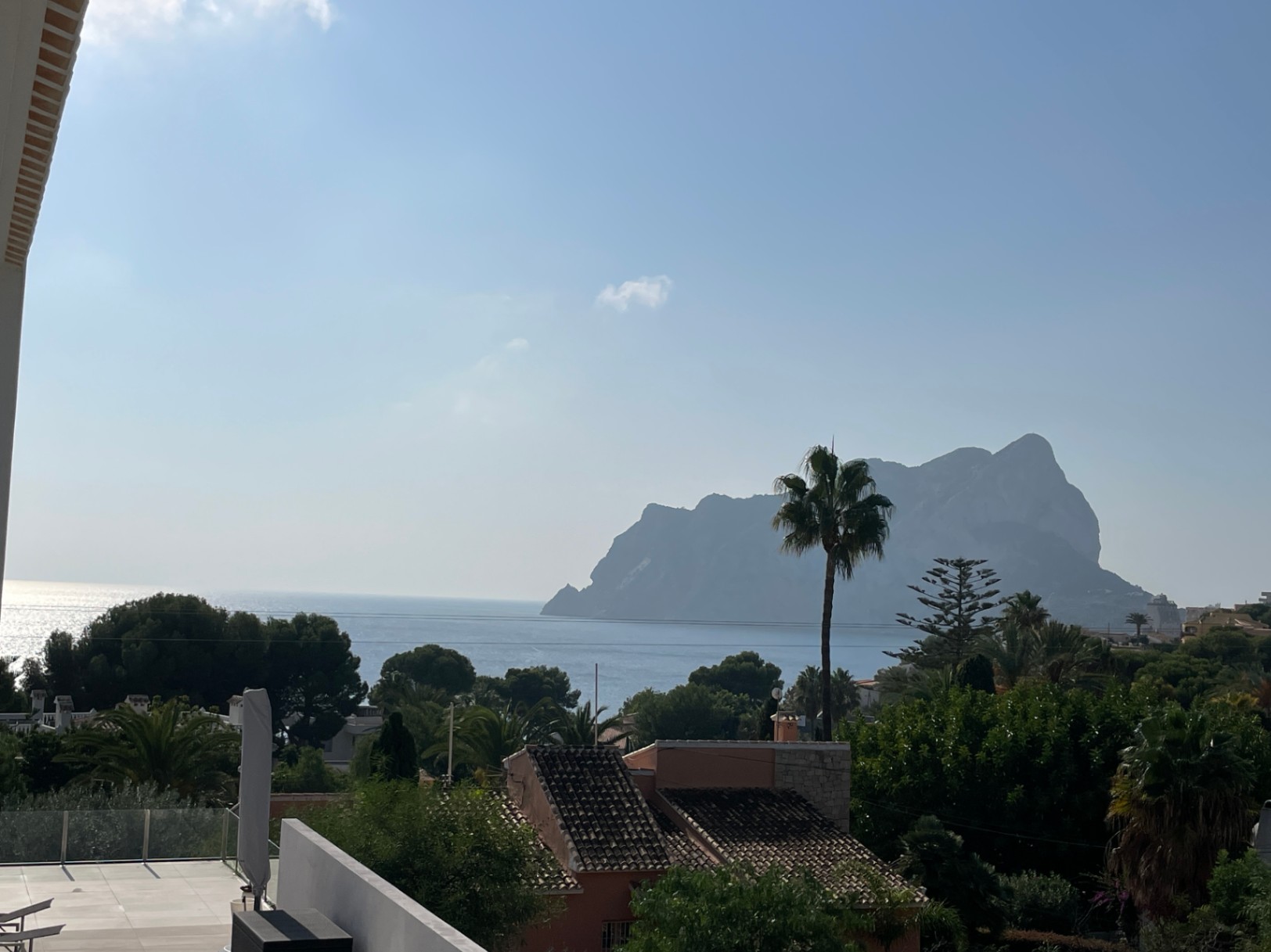 Vue panoramique sur le Peñón de Ifach et la mer Méditerranée depuis une terrasse surélevée avec garde-corps en verre et végétation côtière.