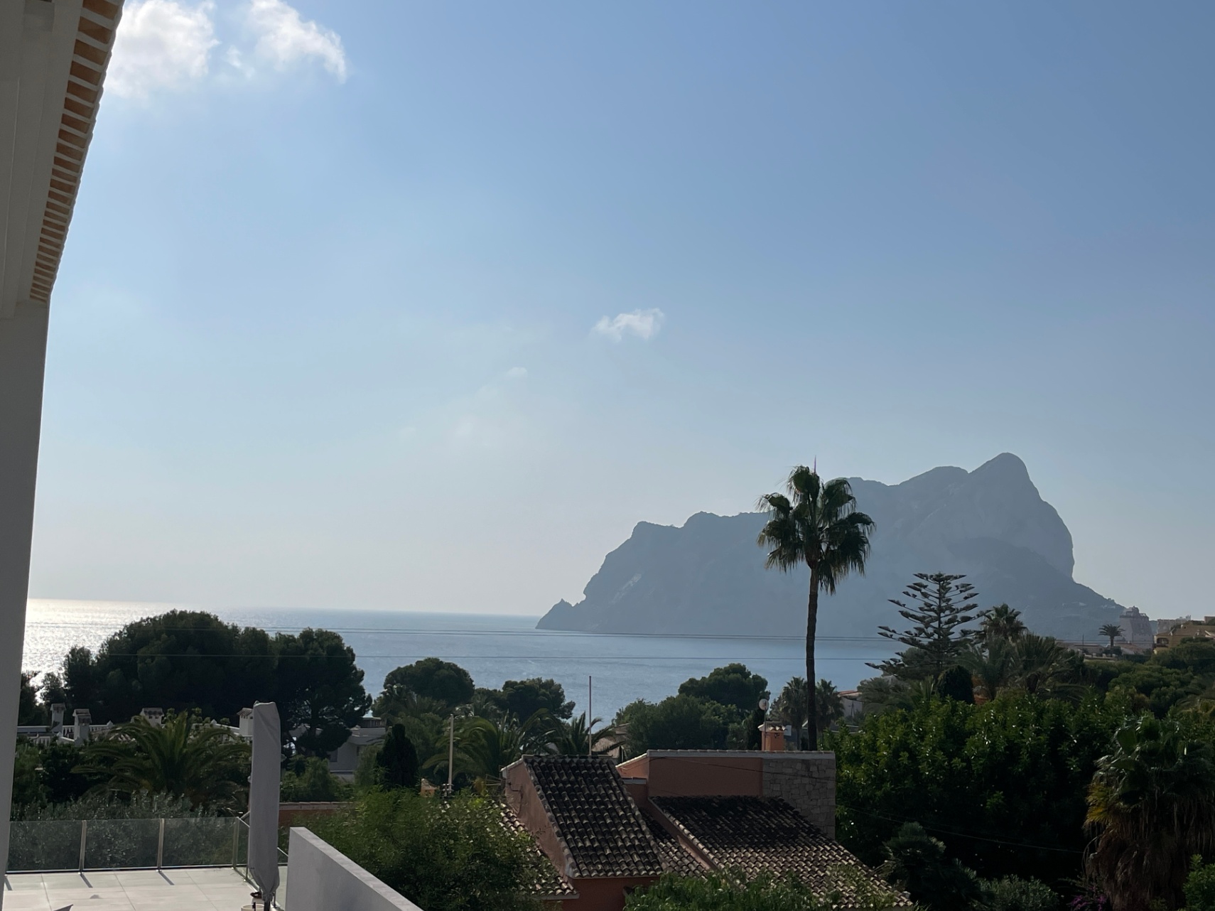 Vues panoramiques sur la mer Méditerranée et le Peñón de Ifach depuis une terrasse surélevée avec garde-corps en verre et végétation luxuriante.