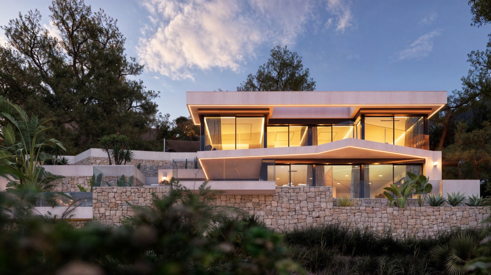 Façade moderne sur deux niveaux avec murs en pierre naturelle, grandes baies vitrées et éclairage LED intégré soulignant les lignes architecturales au crépuscule.