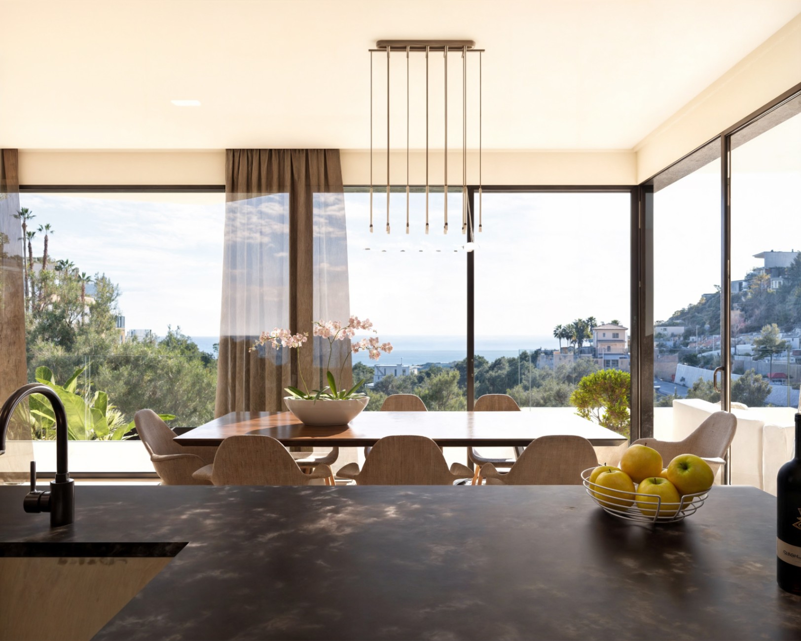 Salle à manger à aire ouverte avec table en bois et baies vitrées offrant une vue sur la mer, vue depuis un plan de travail de cuisine texturé sombre.