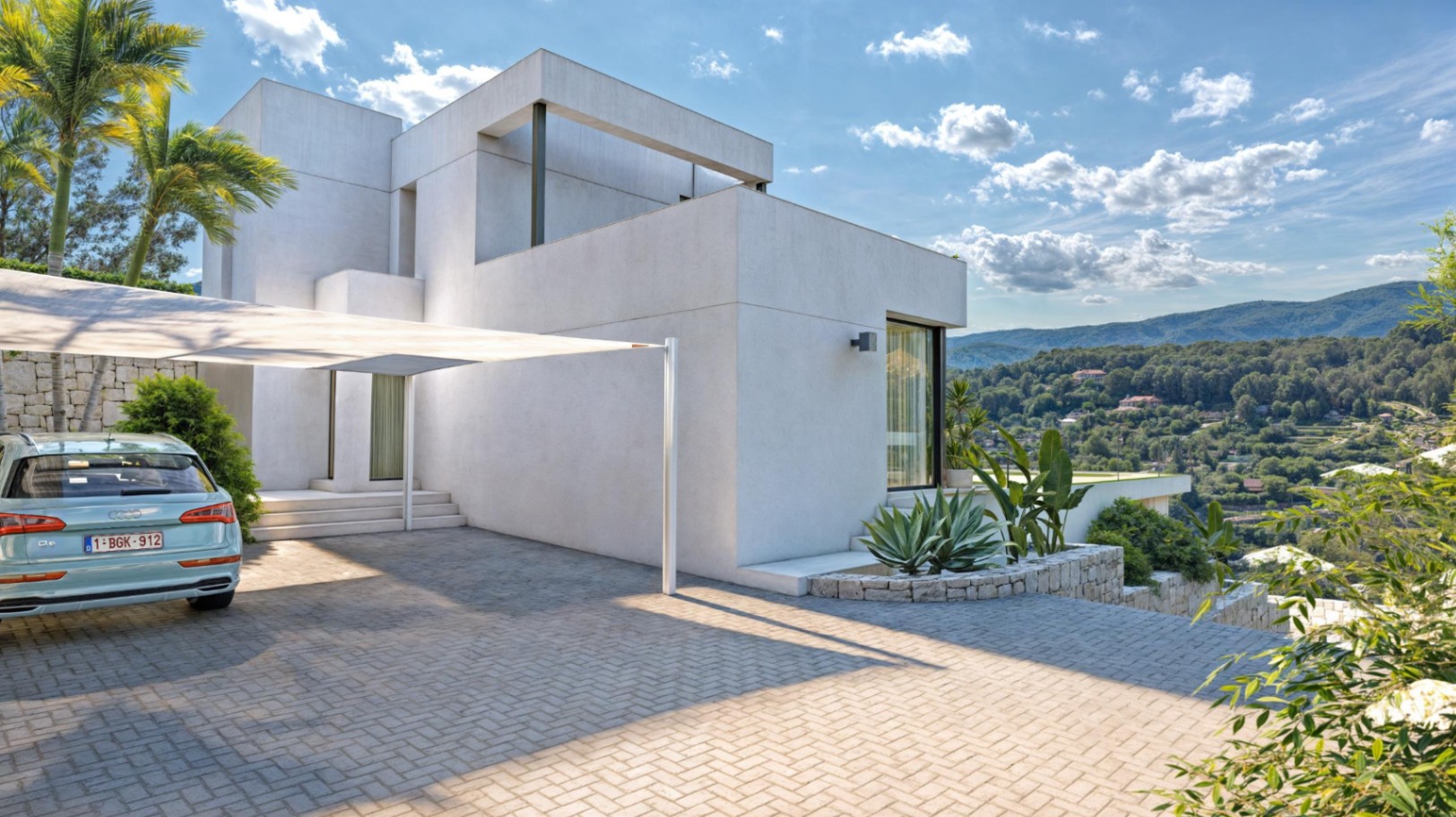 Façade extérieure au design cubique avec parking pavé, voile d'ombrage et murs en pierre naturelle offrant une vue dégagée sur la Sierra de Bernia.