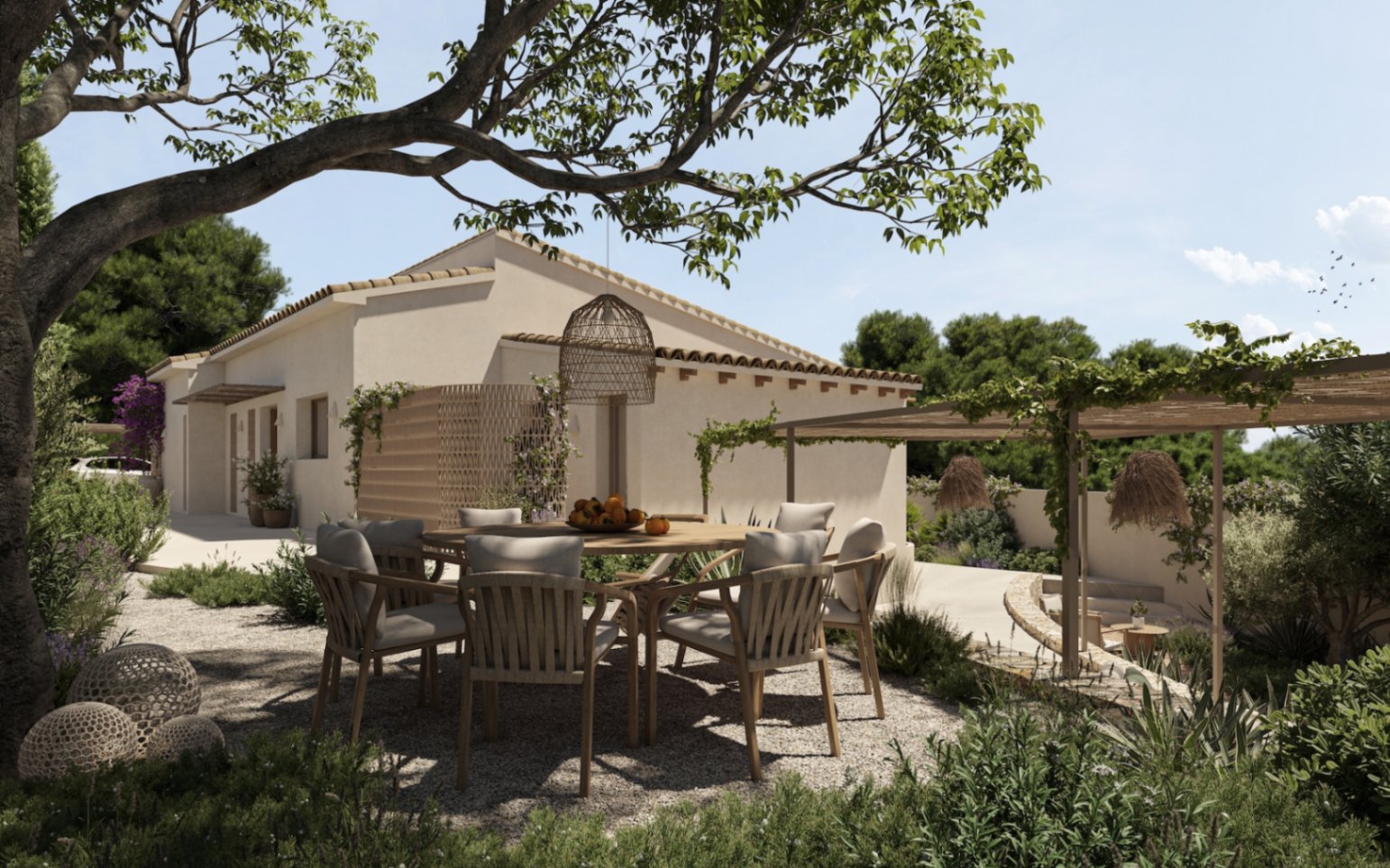 Espace repas extérieur ombragé par un grand arbre, avec table ronde en bois et chaises tressées sur un sol en gravier, bordé de végétation méditerranéenne.