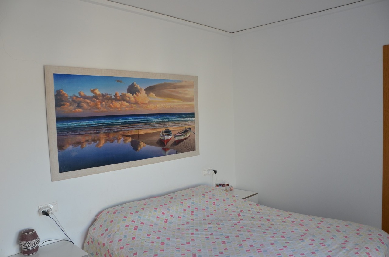 Chambre à coucher avec un grand tableau de paysage côtier au-dessus du lit, des murs blancs et un couvre-lit aux motifs géométriques.