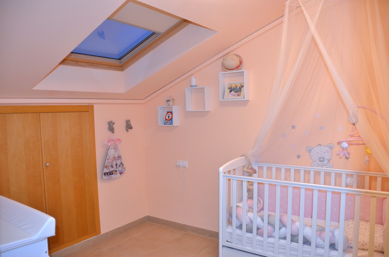 Chambre d'enfant mansardée avec fenêtre de toit, berceau blanc à baldaquin, murs roses et armoire encastrée en bois.