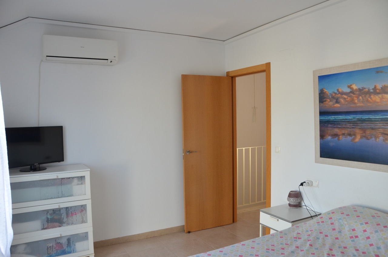 Chambre équipée d'une climatisation murale, d'une commode blanche avec téléviseur et d'une porte en bois ouvrant sur le palier. Sols en grès et style épuré.