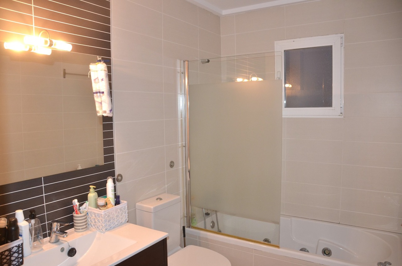 Salle de bain moderne avec meuble vasque blanc, baignoire avec paroi en verre dépoli et carrelage beige complété par un mur d'accent sombre derrière le miroir.