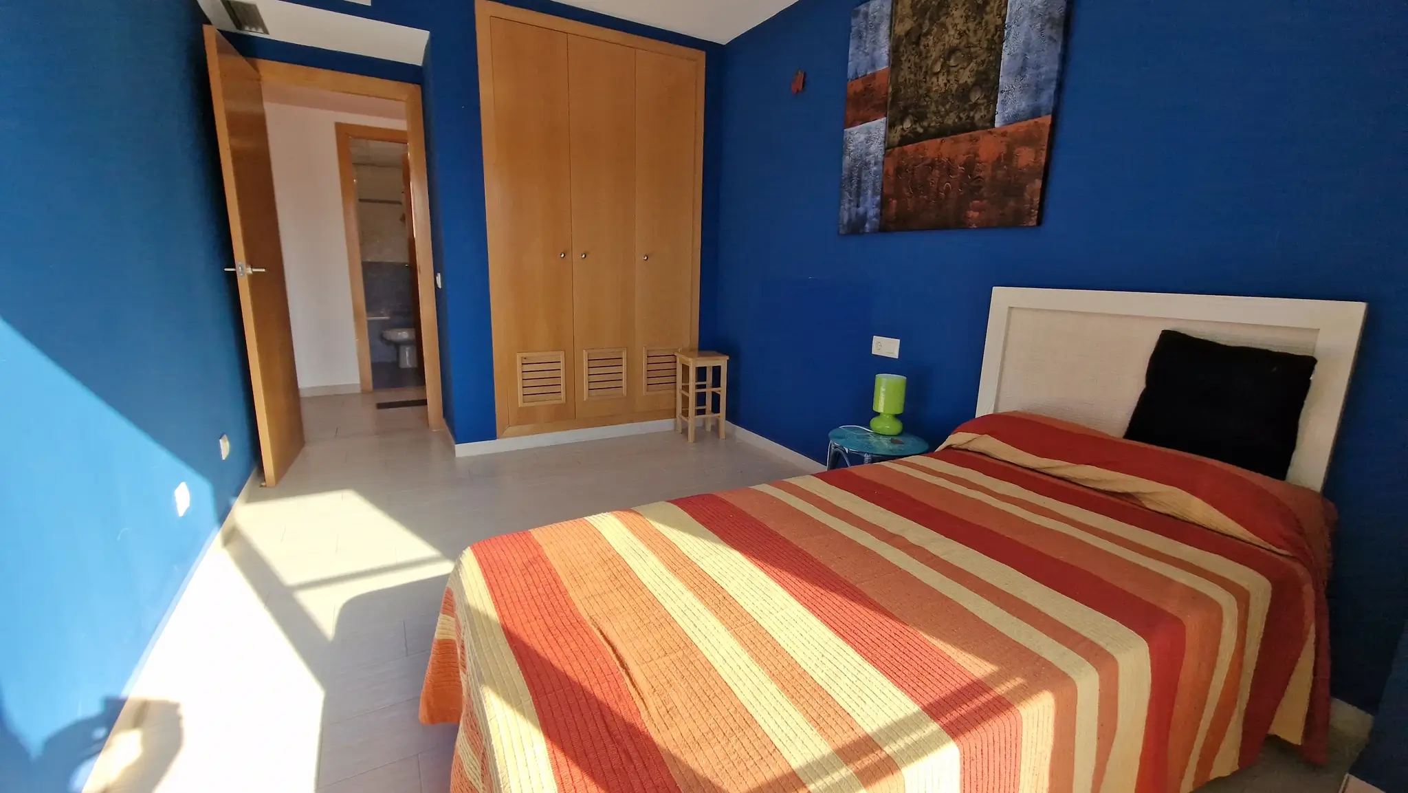 Chambre aux murs bleu profond avec armoire encastrée en bois et sol carrelé. L'espace comprend un lit simple et offre un accès direct à une salle de bain attenante.